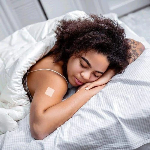 A young woman with curly hair sleeps peacefully on a bed, resting her head on her arms, with a small bandage on her upper arm, surrounded by soft white bedding in a bright room.