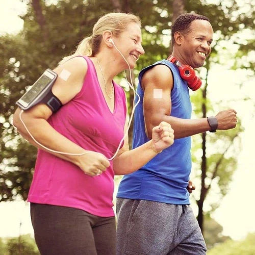A woman in a pink tank top and a man in a blue shirt jog together in a park, both smiling and wearing headphones, enjoying their workout with fitness devices on their arms.