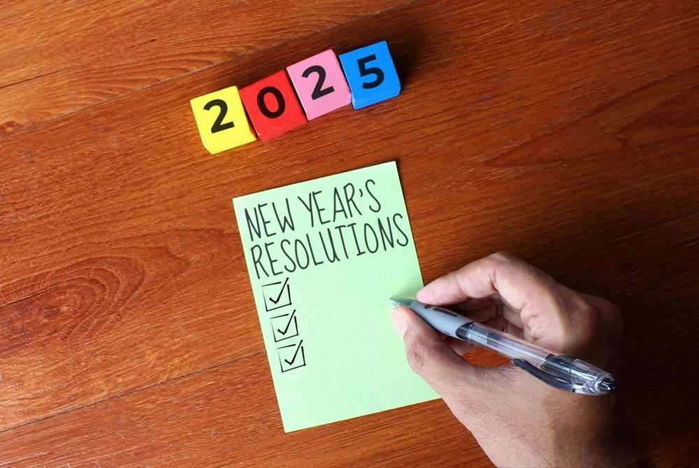 A hand holds a pen writing on a green sticky note labeled "New Year's Resolutions," with three checkboxes marked, and colorful blocks reading "2025" positioned above on a wooden surface.