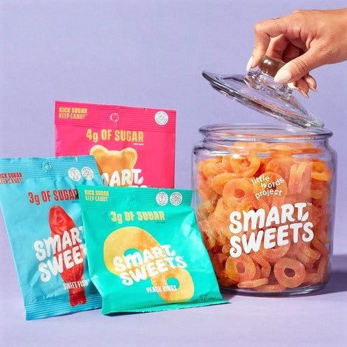 A hand lifts the lid off a glass jar filled with colorful gummy candies labeled "Smart Sweets," alongside two brightly packaged snack bags highlighting low sugar content.
