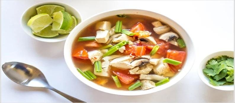 A bowl of chicken soup filled with pieces of chicken, tomatoes, mushrooms, and green onions, accompanied by lime wedges and fresh cilantro on the side.
