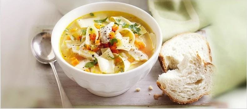 A white bowl filled with chicken vegetable soup featuring noodles, colorful vegetables, and herbs sits on a wooden surface next to a slice of crusty bread and a spoon.