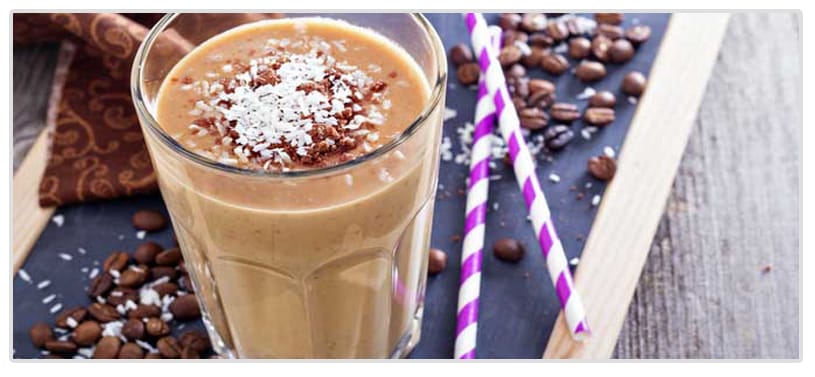 A tall glass of chocolate-coconut shake topped with shredded coconut and cocoa sits on a wooden surface, surrounded by coffee beans and two striped straws.