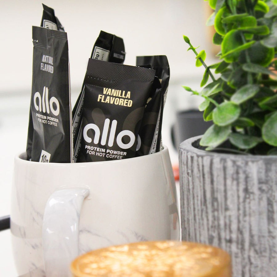 Black packets of Allo protein powder, labeled "Natural" and "Vanilla Flavored," are displayed in a white marble mug next to a textured gray planter and a blurred coffee cup in a bright setting.