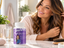 A woman with long, wavy hair smiles while gently touching her hair at a vanity, with a bottle of 