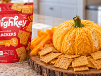 Pumpkin-shaped cheddar cheese ball served with HighKey Cheddar Almond Flour Crackers and fresh orange bell pepper slices on a rustic wooden board, with a hand holding a cracker topped with creamy cheese.