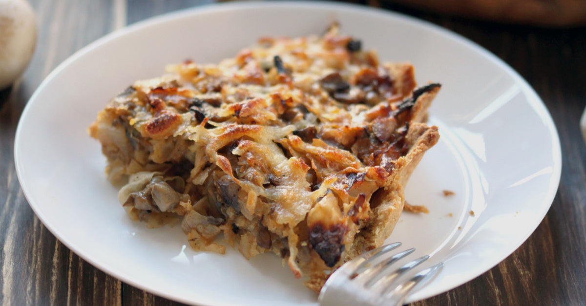 A slice of baked cabbage dish is served on a white plate with a fork beside it, showcasing a golden-brown, crispy top and a hearty filling of cabbage and mushrooms against a wooden table background.