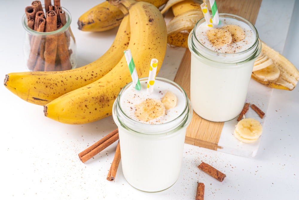 Two glasses of banana smoothie with banana slices and cinnamon on top sit on a wooden cutting board, surrounded by fresh bananas and a jar of cinnamon sticks.
