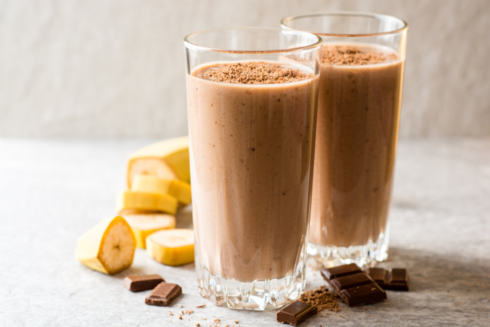 Two tall glasses of chocolate banana smoothie topped with cocoa powder sit on a light surface, surrounded by sliced bananas and pieces of chocolate.
