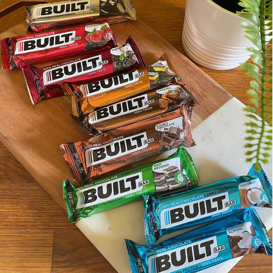 A variety of Built Bars in different flavors are neatly arranged on a wooden cutting board, with a small potted plant visible in the background, showcasing popular protein snack options.