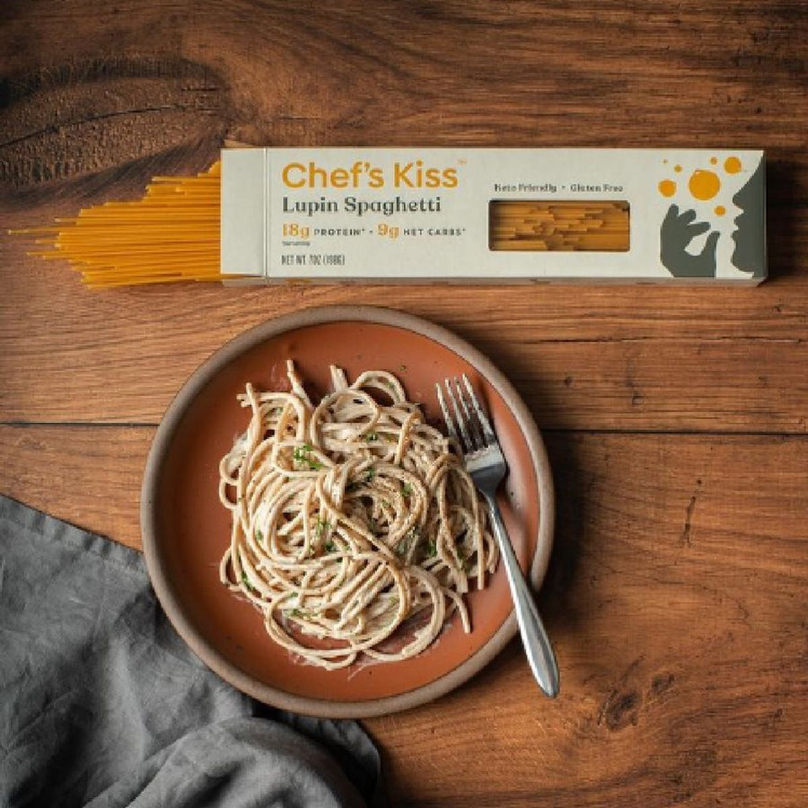 A plate of cooked lupin spaghetti topped with herbs sits next to a box labeled "Chef's Kiss" on a wooden table, with a fork resting on the plate and a gray cloth nearby.