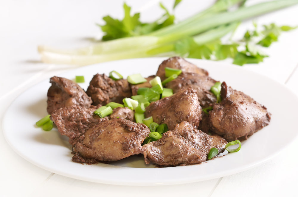 A plate of cooked chicken liver is garnished with chopped green onions, with fresh green herbs in the background, set against a light wooden surface.
