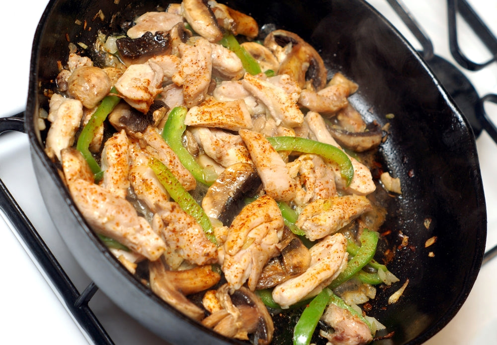 A skillet filled with sautéed chicken strips, green bell peppers, and mushrooms, steaming and seasoned, sits on a stove, showcasing a colorful chicken stir fry dish.