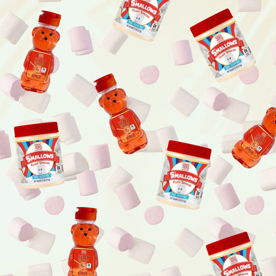 Colorful jars of ChocZero's Smallows Fluff Spread and Hot Honey, shaped like a bear, are surrounded by scattered pink and white marshmallows on a light background.