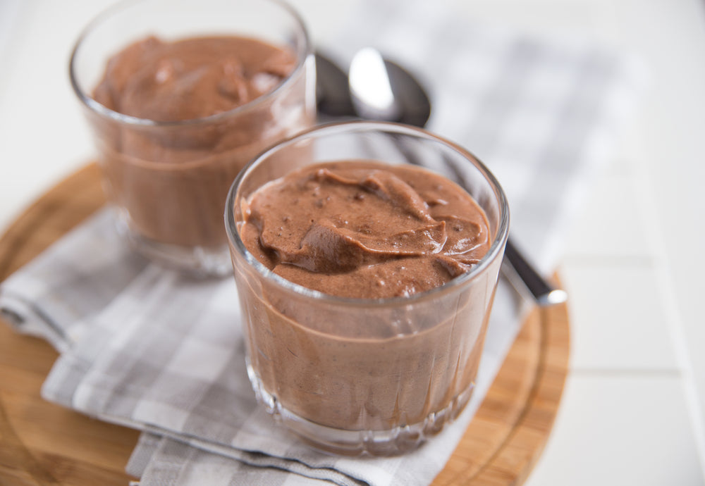 Two glasses of creamy chocolate mousse are placed on a wooden tray, accompanied by a spoon, resting on a checkered napkin against a light background.