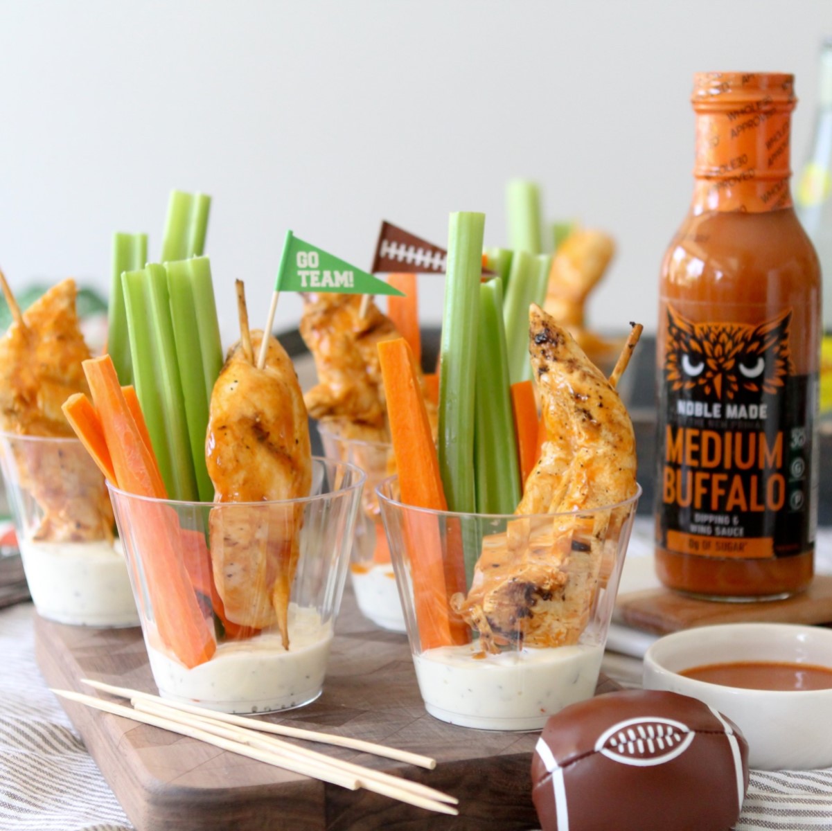 Colorful cups filled with celery, carrot sticks, and grilled chicken skewers are arranged on a wooden board, alongside a bottle of Noble Made Medium Buffalo sauce and a football-shaped snack.