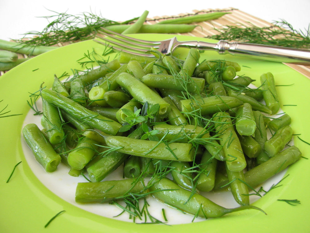 A vibrant green plate holds a generous serving of dilled green beans, garnished with fresh dill, set against a light background with additional green beans in the scene.