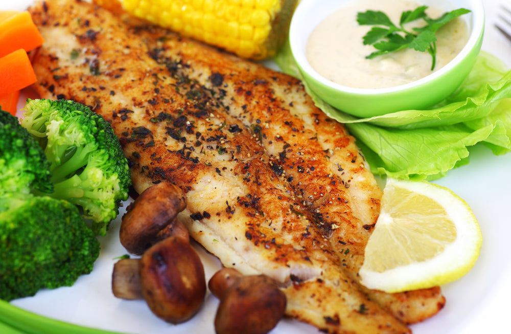 A plate features a golden-brown fish fillet garnished with herbs, accompanied by broccoli, carrots, mushrooms, corn, a wedge of lemon, and a small bowl of sauce topped with cilantro.