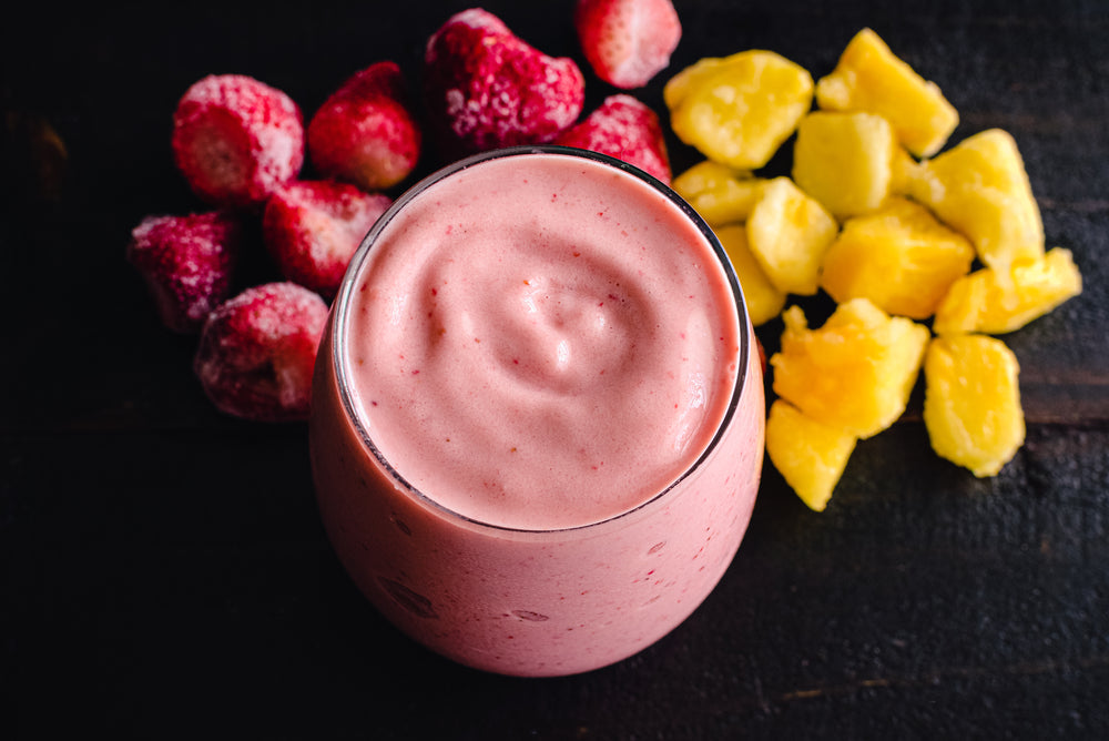 A creamy pink smoothie is centered in a glass, surrounded by frozen strawberries and chunks of yellow pineapple on a dark wooden surface.