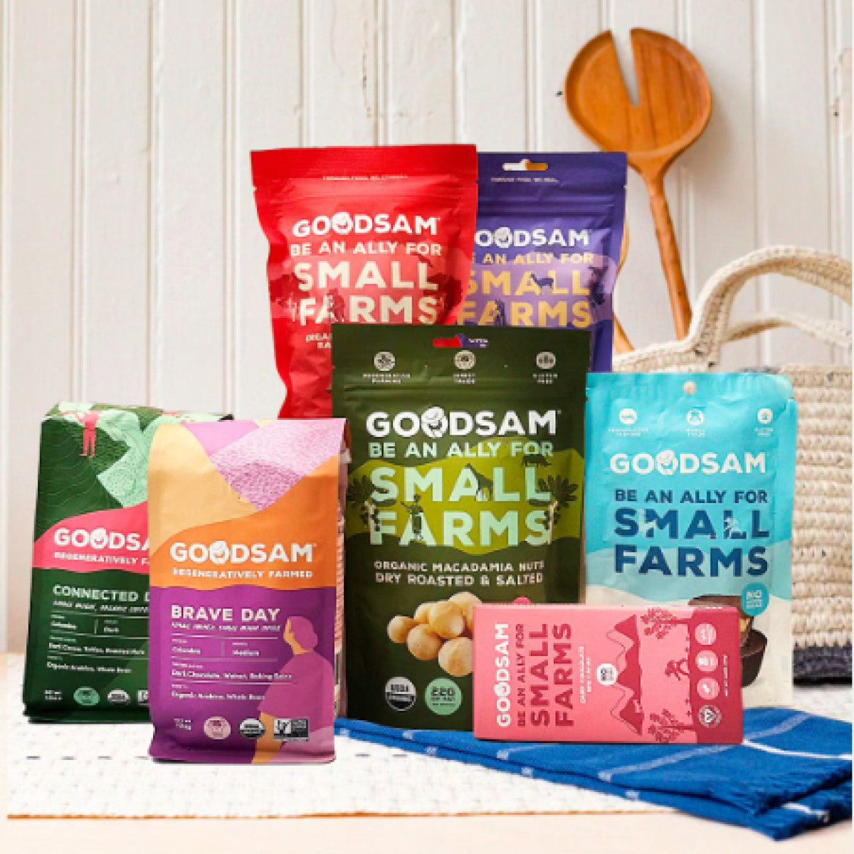 A variety of colorful GoodSam snack packages, including macadamia nuts and other treats, are displayed on a textured surface with a wooden spoon and woven basket in the background.