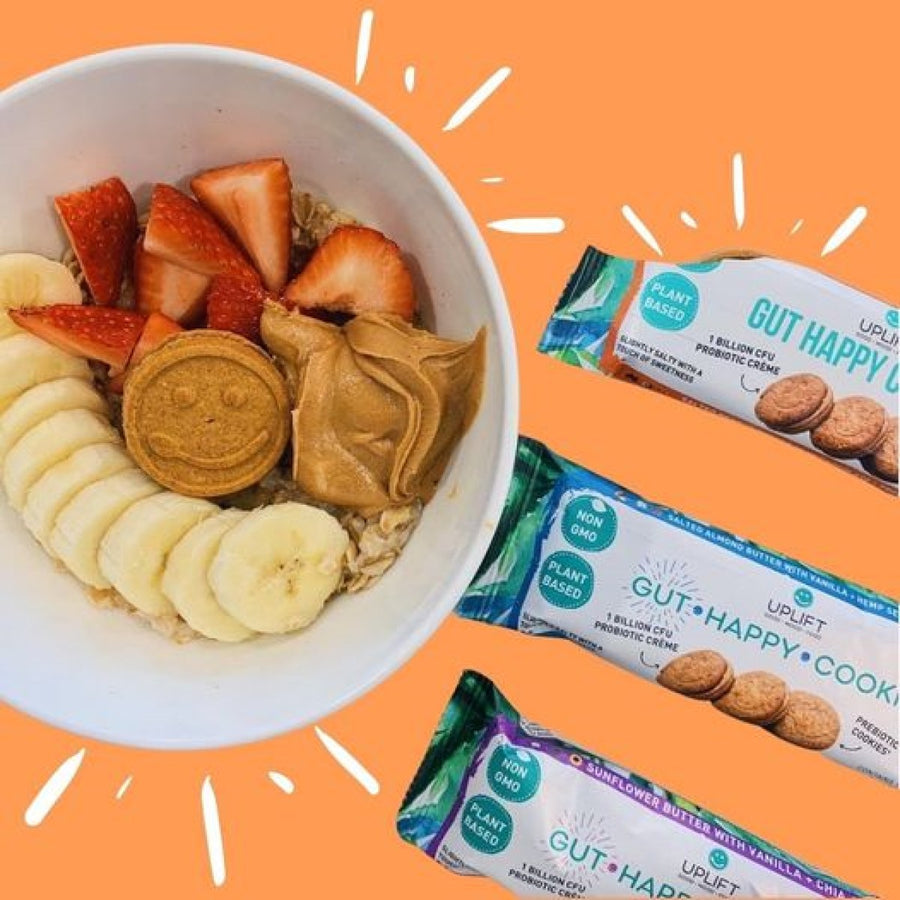 A bowl of oatmeal topped with sliced bananas, strawberries, a smiling cookie, and peanut butter is shown alongside Uplift Food Gut Happy Cookies against a bright orange background.