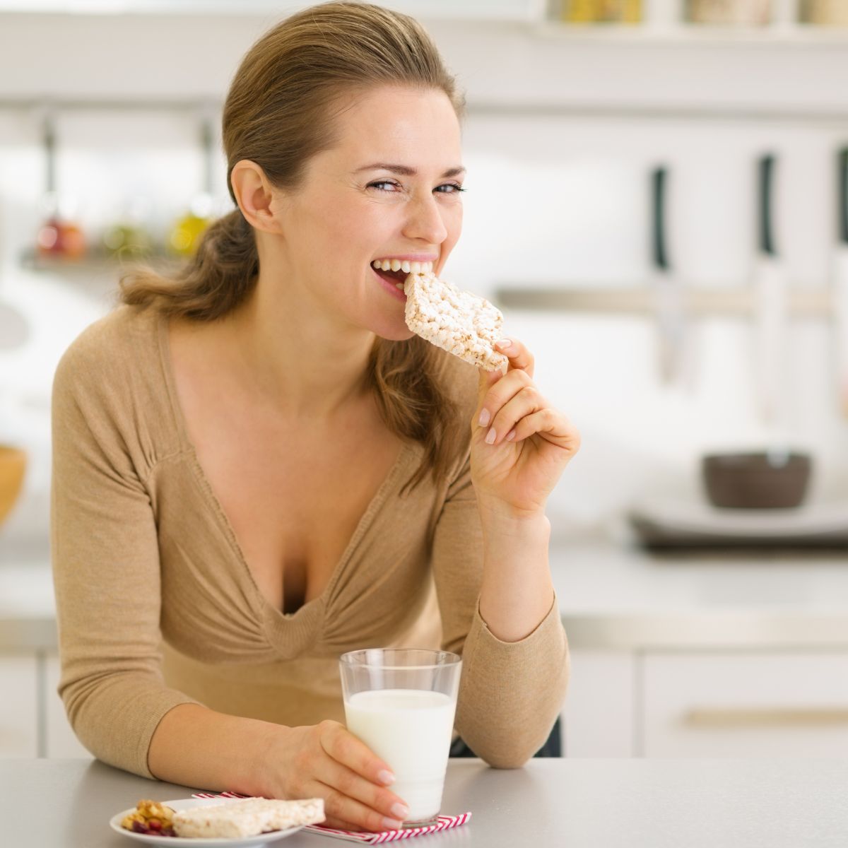 A woman smiles while taking a bite of a rice cake, holding a glass of milk in her other hand, seated at a kitchen table with snacks and a bright, modern kitchen in the background.