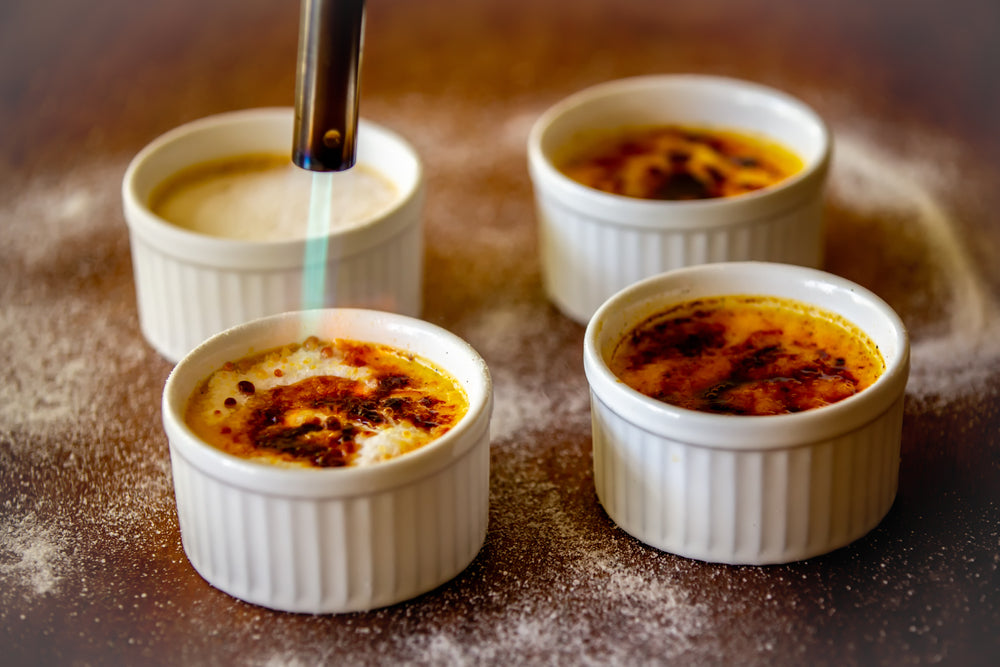 A kitchen setting features four white ramekins with golden-brown tops, as a kitchen torch caramelizes the sugar on one, surrounded by a dusting of powdered sugar.