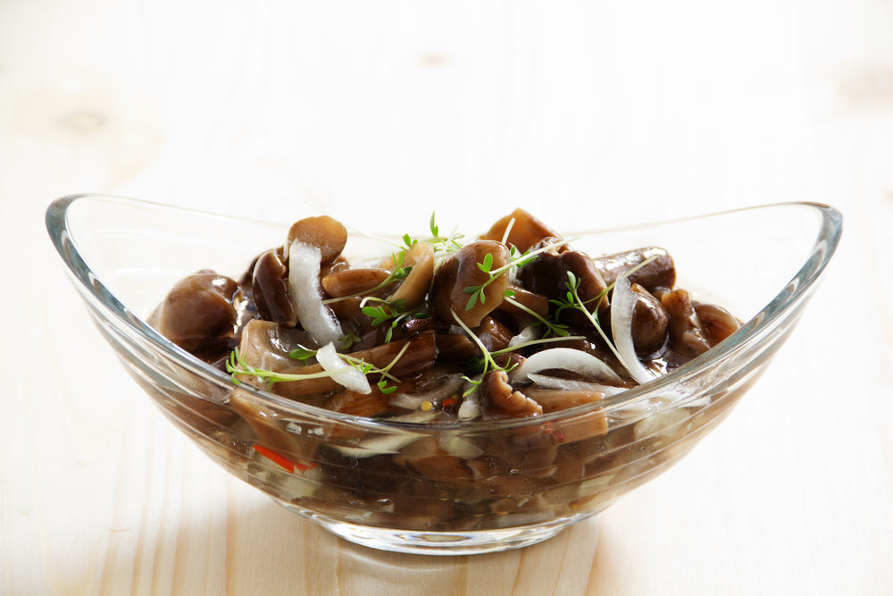 A glass bowl filled with marinated mushrooms, garnished with thin slices of onion and fresh herbs, sits on a light wooden surface, showcasing a savory dish ready to be served.
