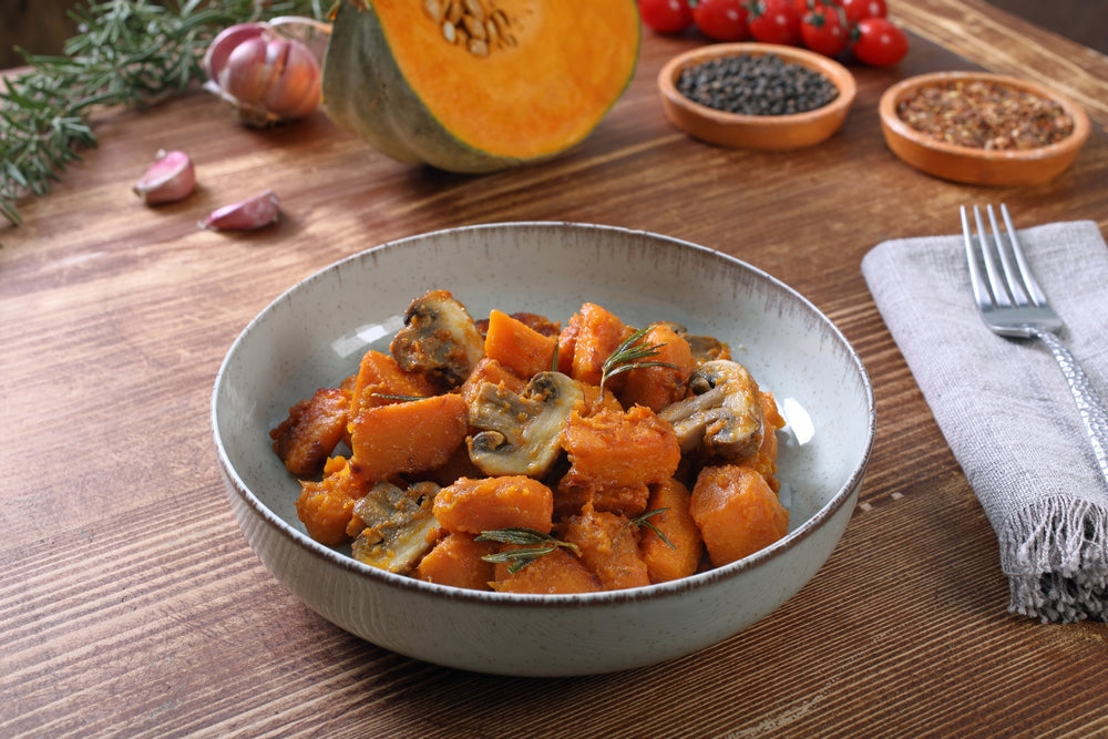 A bowl of roasted butternut squash and mushrooms garnished with rosemary sits on a wooden table, surrounded by garlic, cherry tomatoes, and spices in small bowls.
