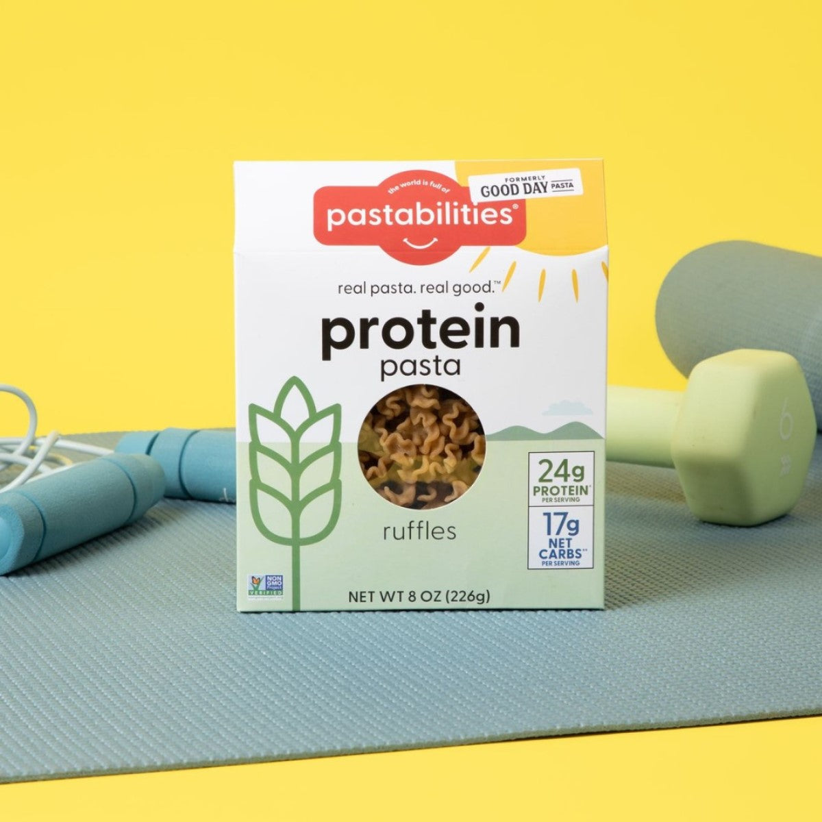 A box of Pastabilities protein pasta labeled "ruffles" sits on a light blue exercise mat, with a jump rope and a green dumbbell nearby, against a bright yellow background.