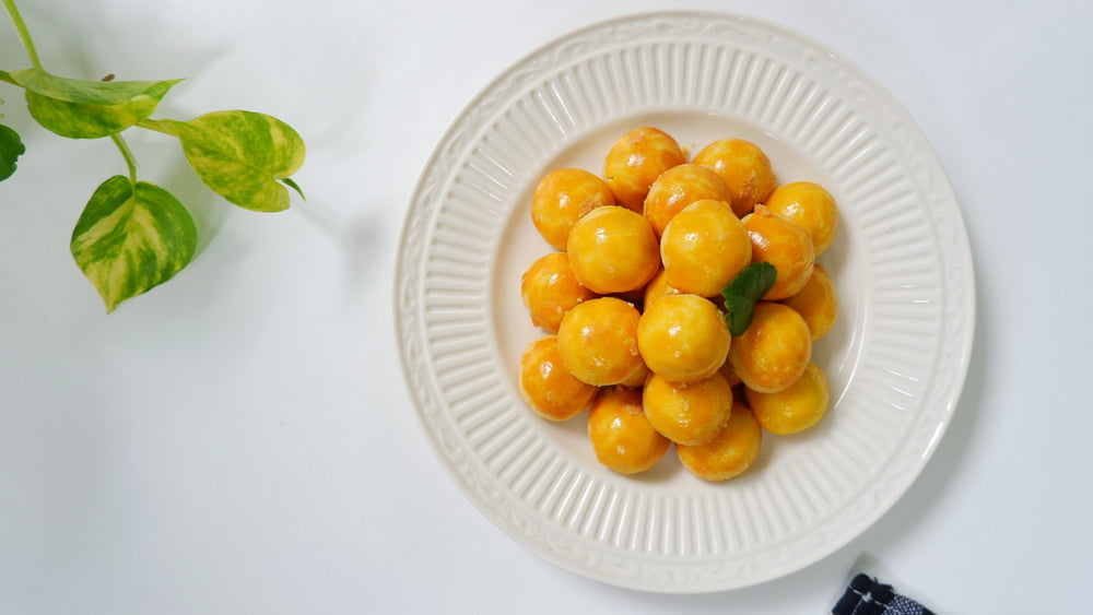 A plate of golden, round cookies resembling Pina Coladas is arranged in a pyramid shape, garnished with a small green leaf, with a plant and a dark cloth visible in the background.