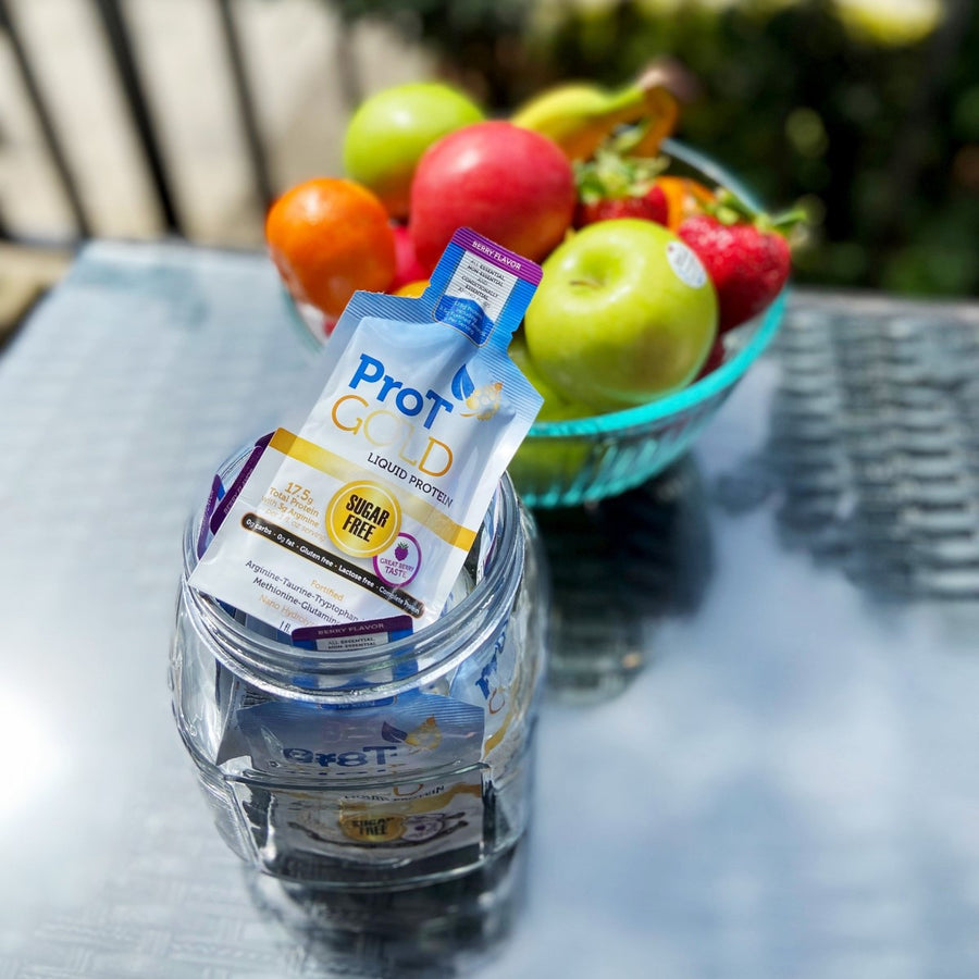 A packet of ProT Gold liquid protein sits in a glass jar on a table, with a bowl of colorful fruits including apples, oranges, and strawberries in the background under bright sunlight.