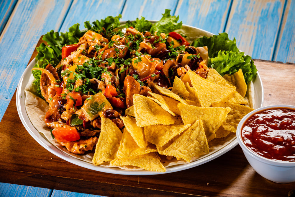 A colorful protein taco salad with seasoned chicken, fresh vegetables, and cheese is served on a bed of lettuce with tortilla chips and a small bowl of salsa on a wooden table.