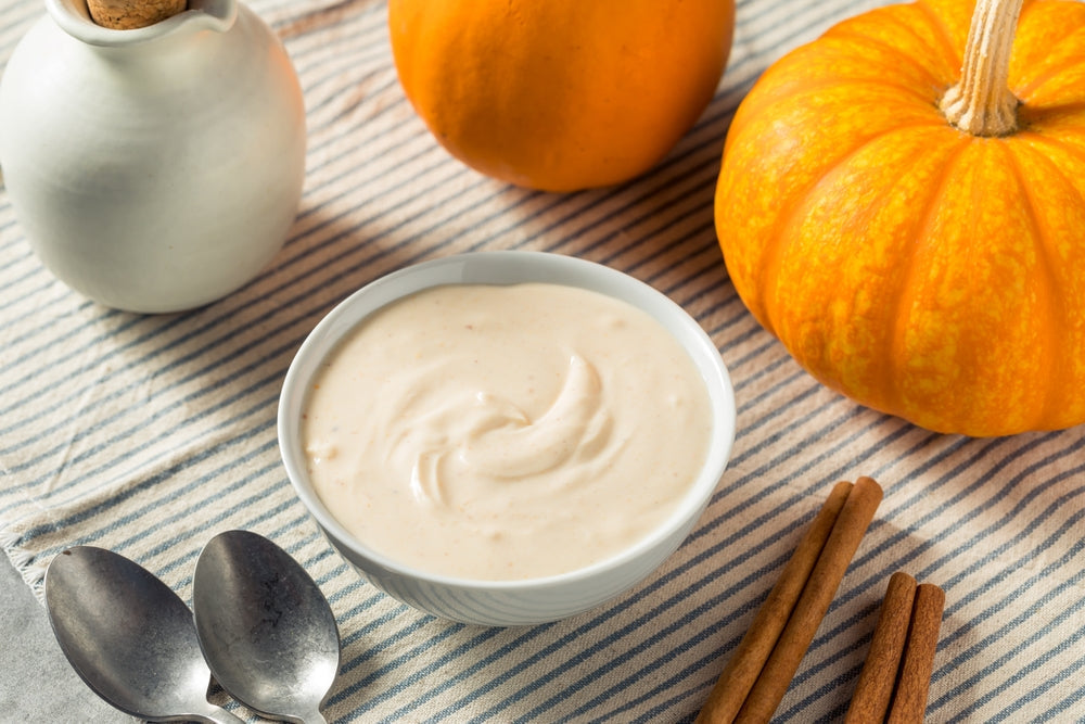 A bowl of creamy pumpkin pie pudding sits on a striped cloth, surrounded by two bright orange pumpkins and cinnamon sticks, suggesting a cozy autumn dessert setting.