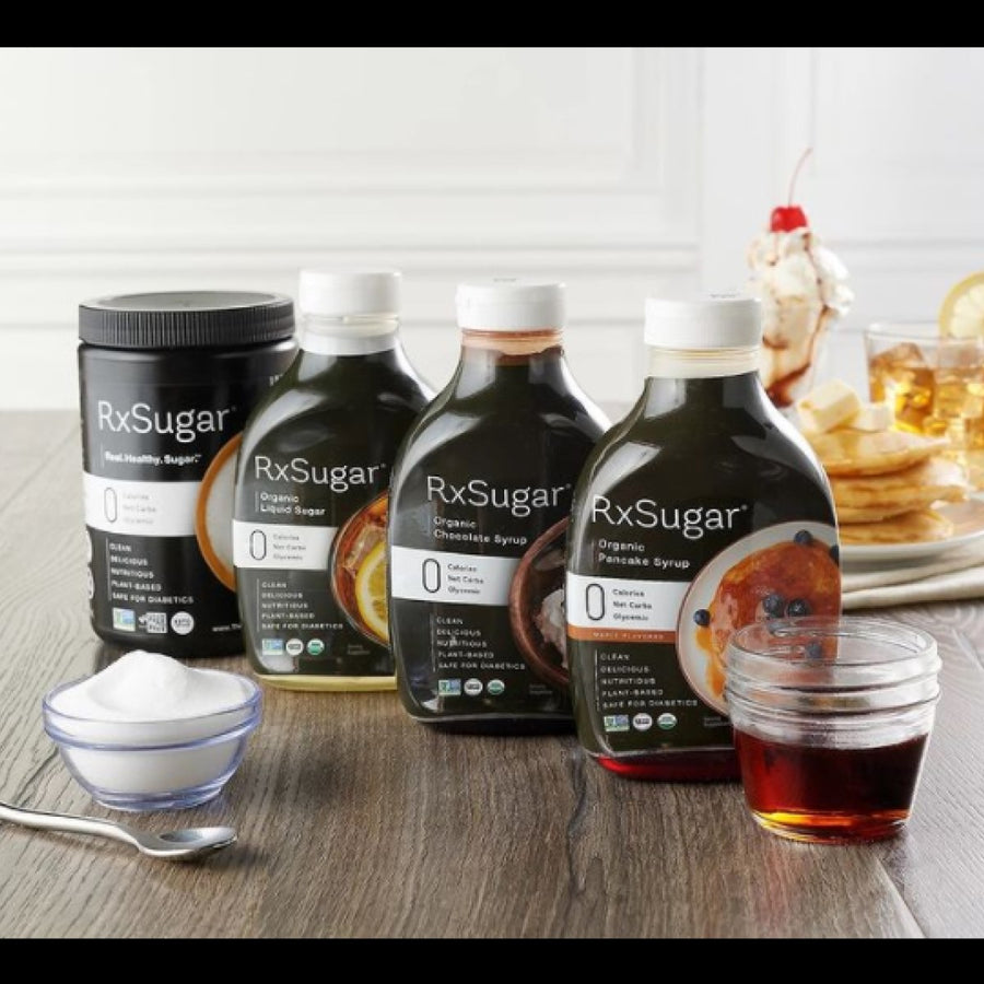 Four bottles of RxSugar organic sweeteners and syrups are displayed on a wooden table, alongside a small bowl of sugar and a glass of syrup, with pancakes in the background.