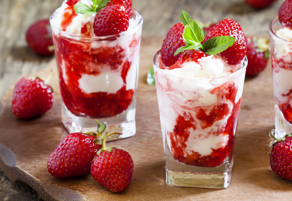 Three clear glasses filled with raspberry slushie layered with cream and topped with fresh strawberries and mint leaves, placed on a wooden surface surrounded by whole strawberries.