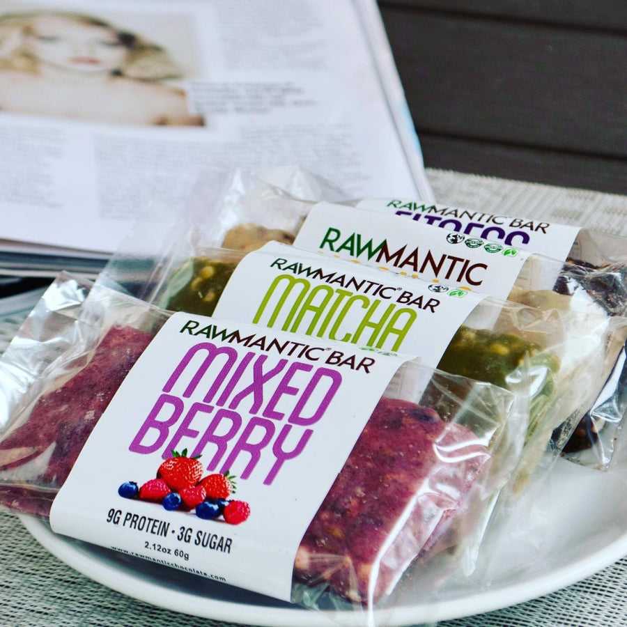 Three Rawmantic Chocolate bars labeled "Mixed Berry," "Matcha," and "Cacao" are displayed on a plate, with a magazine featuring a model in the background, suggesting a relaxed setting.