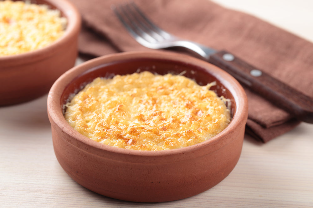 Two small clay bowls of golden-brown rice pudding sit on a light wooden surface, with a fork and brown napkin in the background, showcasing a creamy dessert topped with a crispy layer.