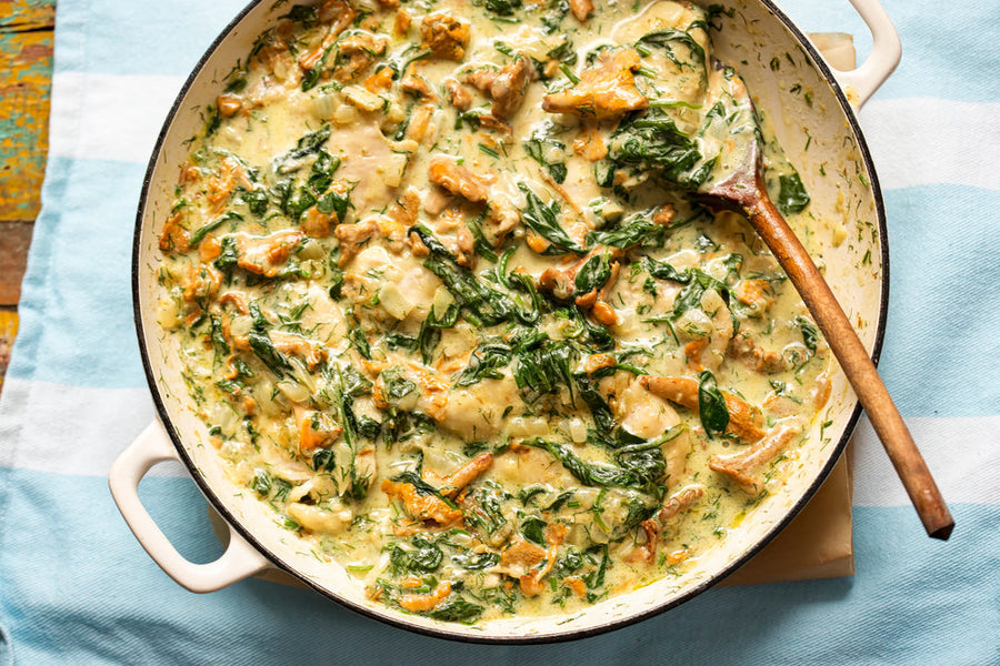A creamy spinach casserole with sautéed mushrooms and herbs is served in a round dish, with a wooden spoon resting inside, placed on a light blue striped cloth.