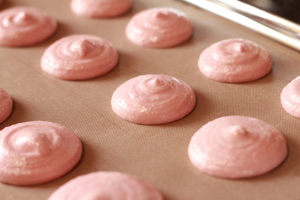 Pale pink meringue dollops are arranged in neat rows on a baking sheet lined with silicone, ready for baking to create strawberry almond cookies.