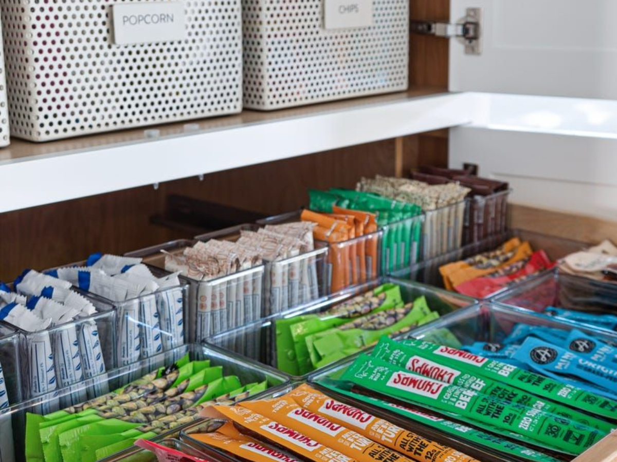A neatly organized pantry shelf displays various sugar-free snacks in clear containers, including protein bars and nuts, alongside labeled bins for popcorn and chips.