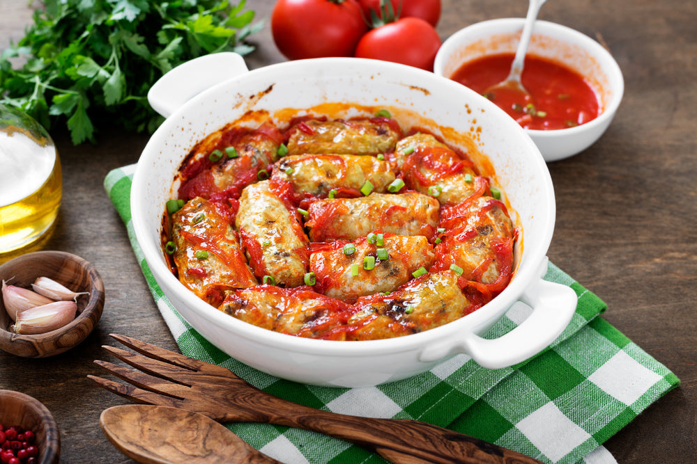 A white bowl filled with sweet cabbage rolls in tomato sauce is placed on a green and white checkered cloth, surrounded by fresh tomatoes, garlic, and a small bowl of sauce.