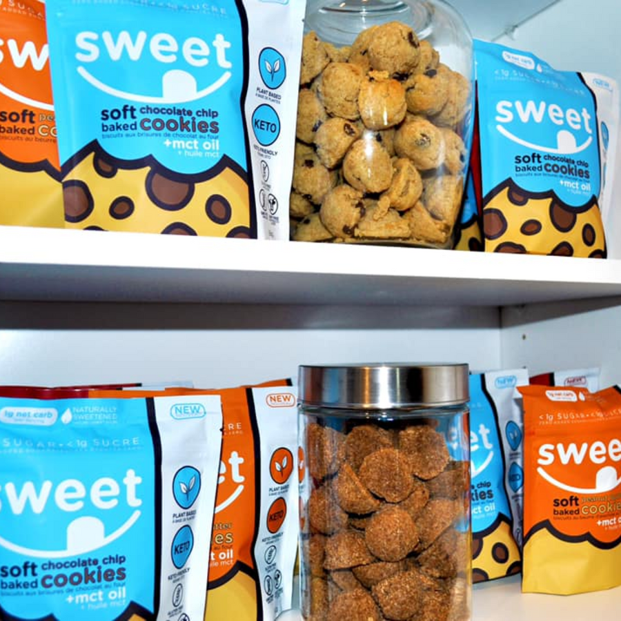 A shelf displays various packages of Sweet Nutrition plant-based low-carb cookies, including soft chocolate chip varieties, alongside a glass jar filled with round cookies.