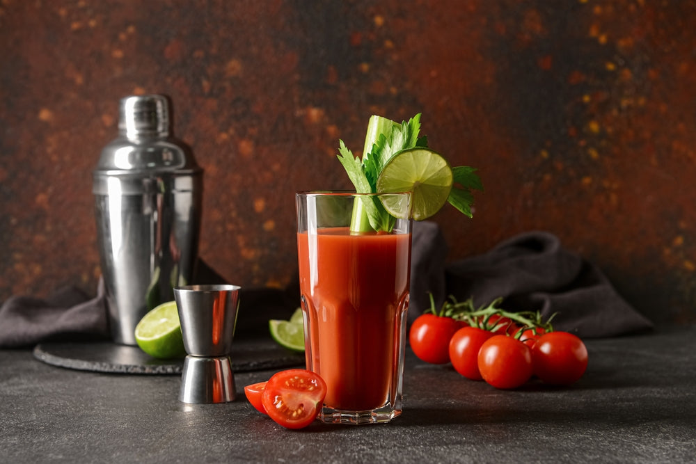 A tall glass of tomato cocktail garnished with celery and lime sits on a dark surface, accompanied by a cocktail shaker and fresh tomatoes, against a textured, warm-colored background.