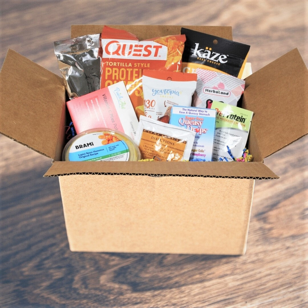 A cardboard box is open and filled with various health and protein snack products, including protein chips, supplements, and snacks, set against a wooden background.
