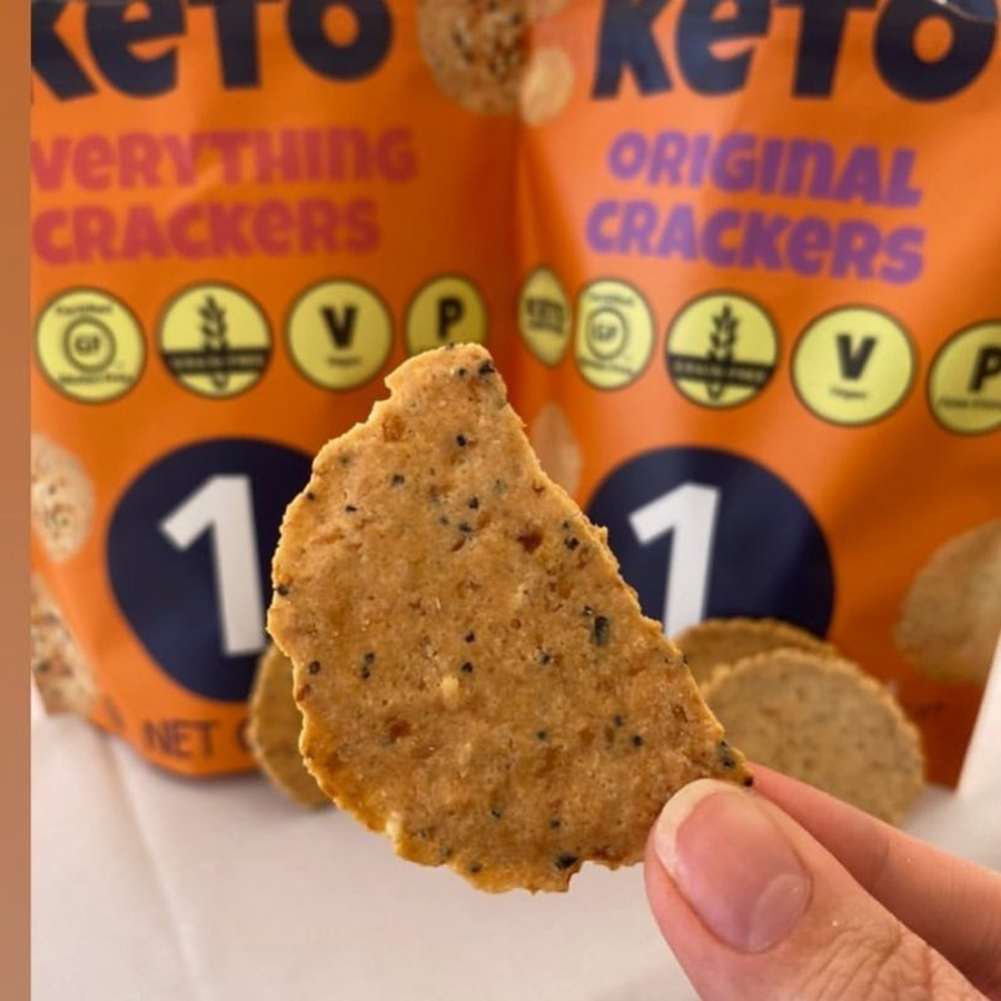 A hand holds a piece of a keto cracker in front of two colorful bags labeled "Keto Everything Crackers" and "Keto Original Crackers," showcasing their low-carb snack options.