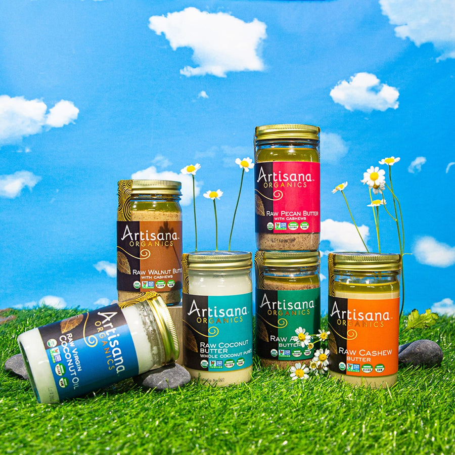 Six jars of Artisana organic nut butters and coconut oil are arranged on green grass with small white flowers, set against a bright blue sky with fluffy clouds.