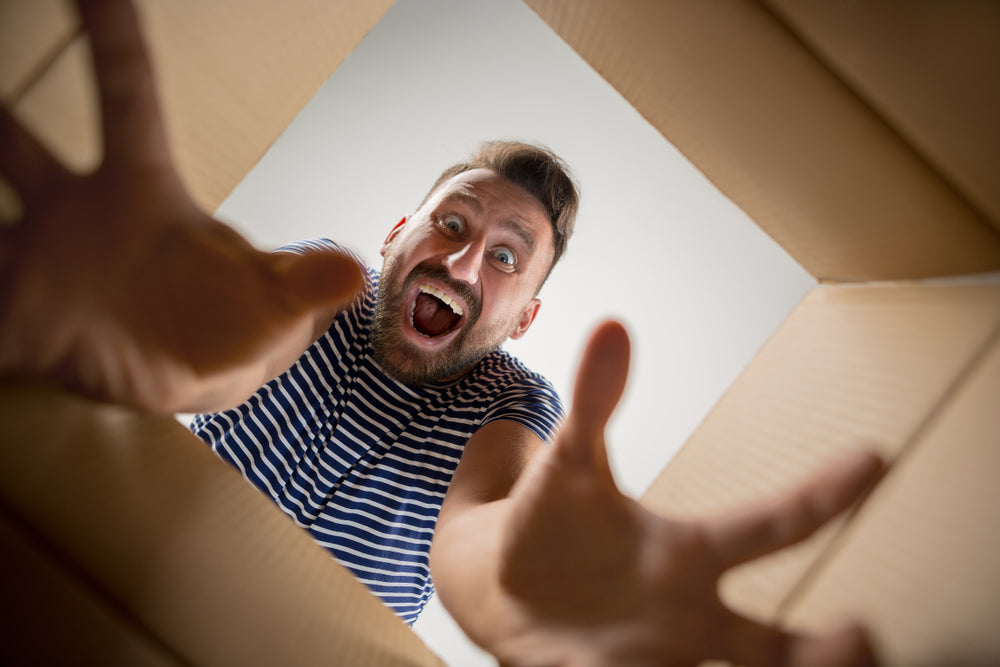 A man with a surprised expression reaches into a cardboard box, looking excited as he discovers its contents, set against a plain background.