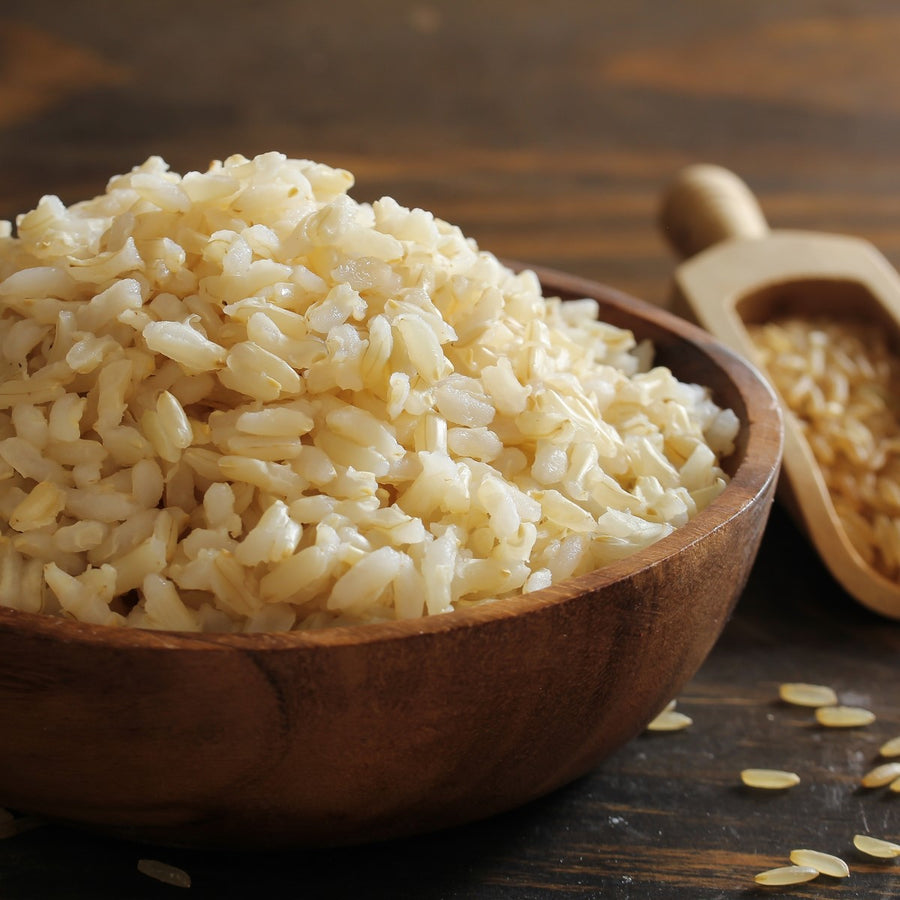 A wooden bowl filled with cooked brown rice sits on a wooden surface, with a small scoop nearby containing more rice grains, highlighting the texture and color of the rice.