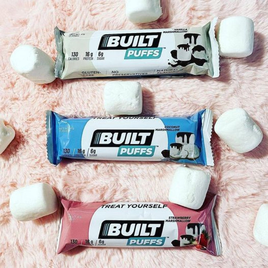 Three Built Bar Protein Puffs in vanilla, coconut, and strawberry flavors are displayed on a soft pink surface, surrounded by fluffy marshmallows.